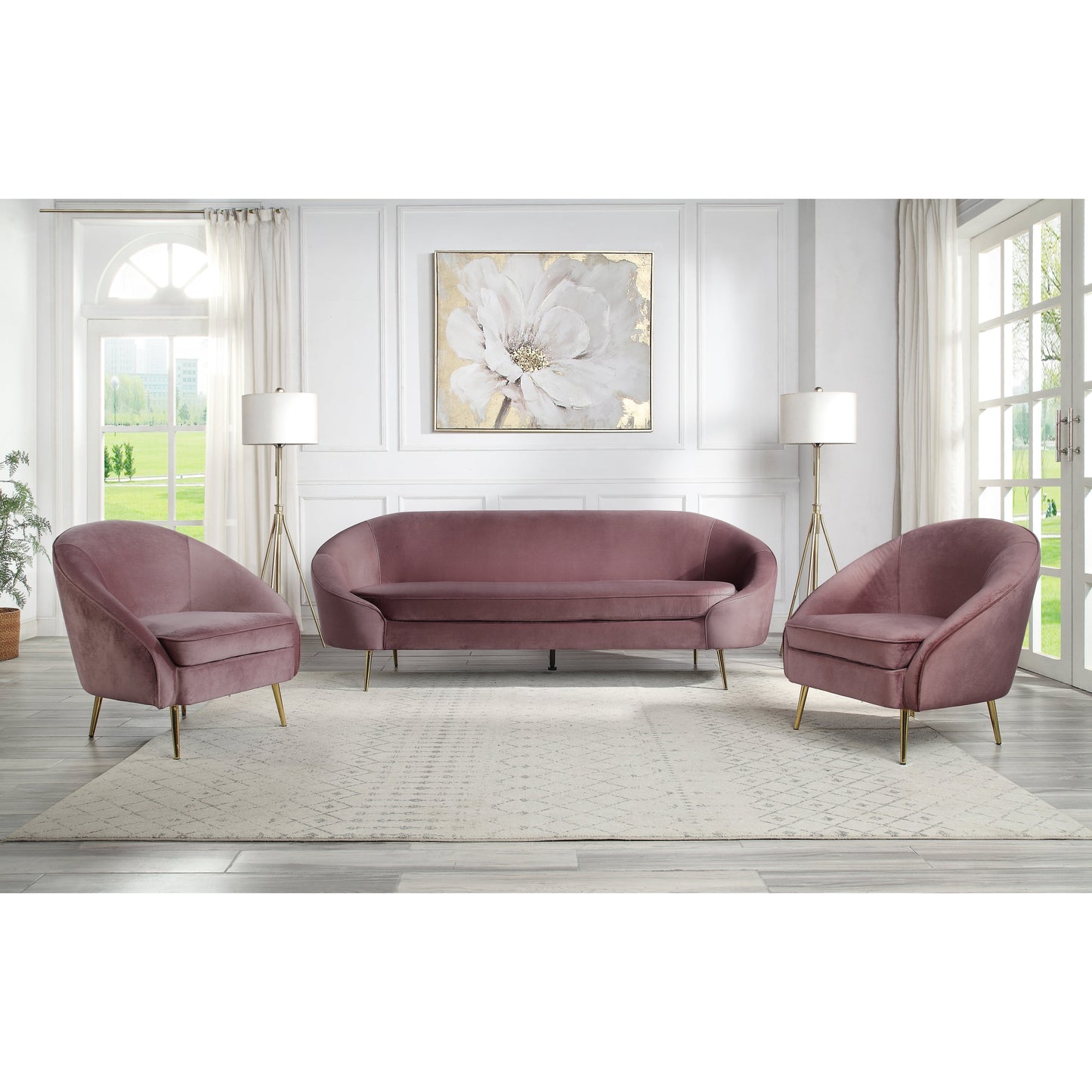 Decorated living room showing the Abey sofa set, which includes a sofa and accent chair. The Abey living room collection is not only visually striking, but it also offers plenty of comfort. The curved silhouette creates a glam appearance. Upholstered in a luxurious mauve velvet fabric to add dimension to the pieces. The shiny gold metal tapered legs add another pop color and emphasize the glamorous design. This set provides enough seating for all your guests.